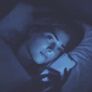 Lumière bleue et sommeil : quel impact réel ? Femme dans son lit regardant un smartphone éclairé en bleu, illustrant l'impact de la lumière bleue sur le sommeil.