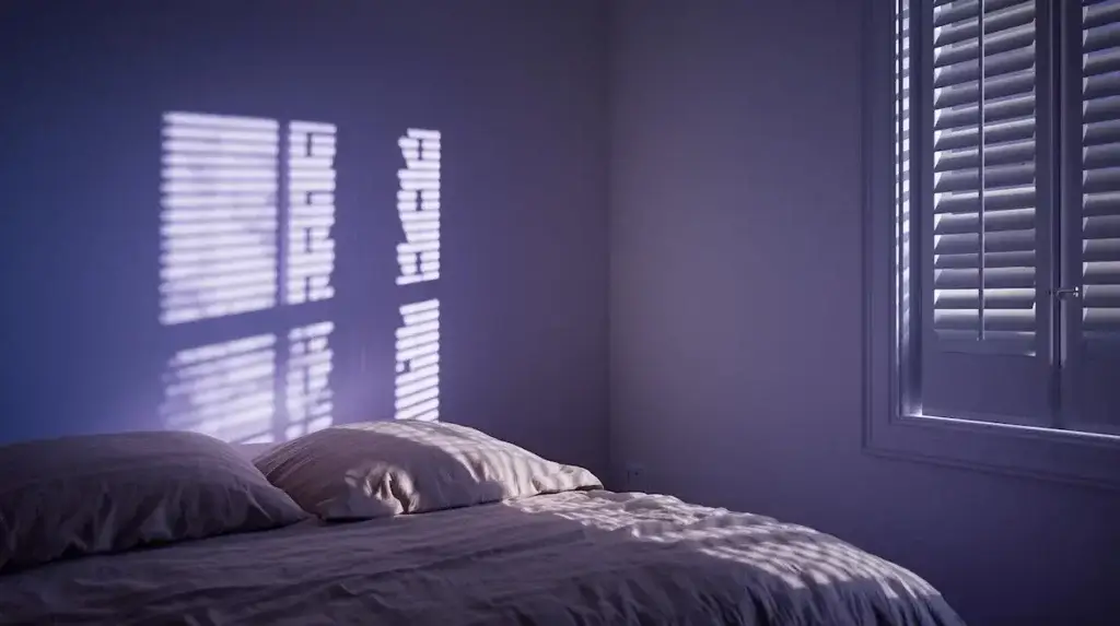 Chambre minimaliste au crépuscule avec lumière tamisée filtrant par des volets, illustrant l'obscurité nécessaire pour lutter contre la pollution lumineuse et améliorer le sommeil