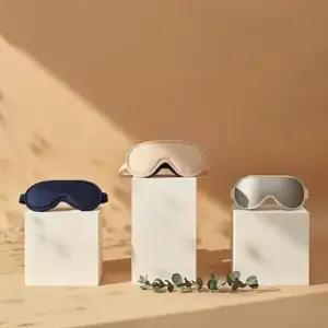 Meilleur masque de sommeil : Top 10 pour mieux dormir en 2026 Trois masques de sommeil premium disposés sur podium à hauteurs différentes : masque soie bleu marine, masque 3D ergonomique beige sur socle central plus haut, et masque connecté argenté, avec eucalyptus au premier plan
