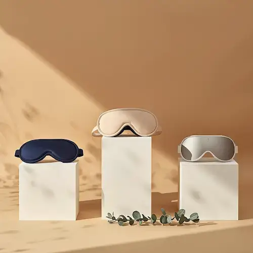 Trois masques de sommeil premium disposés sur podium à hauteurs différentes : masque soie bleu marine, masque 3D ergonomique beige sur socle central plus haut, et masque connecté argenté, avec eucalyptus au premier plan