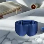 Olesilk : avis sur le masque de sommeil en soie (test 2025) Masque de sommeil Olesilk bleu nuit en soie, présenté sur un fond blanc marbré avec un livre ouvert et une bougie floue en arrière-plan.