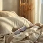 Manta Sleep Mask : test, avis et comparatif du masque de sommeil iconique Manta Sleep Mask gris posé sur lit avec draps lin blanc cassé, lumière naturelle dorée de matin, ambiance chambre sereine