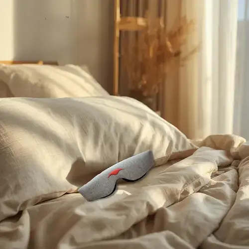 Manta Sleep Mask gris posé sur lit avec draps lin blanc cassé, lumière naturelle dorée de matin, ambiance chambre sereine
