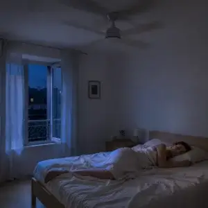 Ventilateur plafond silencieux : comment choisir un modèle adapté à une chambre ? Ventilateur de plafond silencieux en marche dans une chambre la nuit, avec une personne dormant sous un drap léger près d’une fenêtre ouverte.