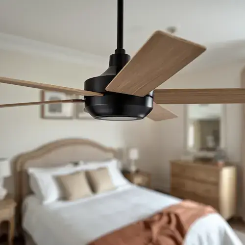 Ventilateur de plafond aux pales en bois clair et armature noire mate, installé dans une chambre cosy aux tons terracotta, illustrant le bon sens de rotation à régler entre l'été et l'hiver